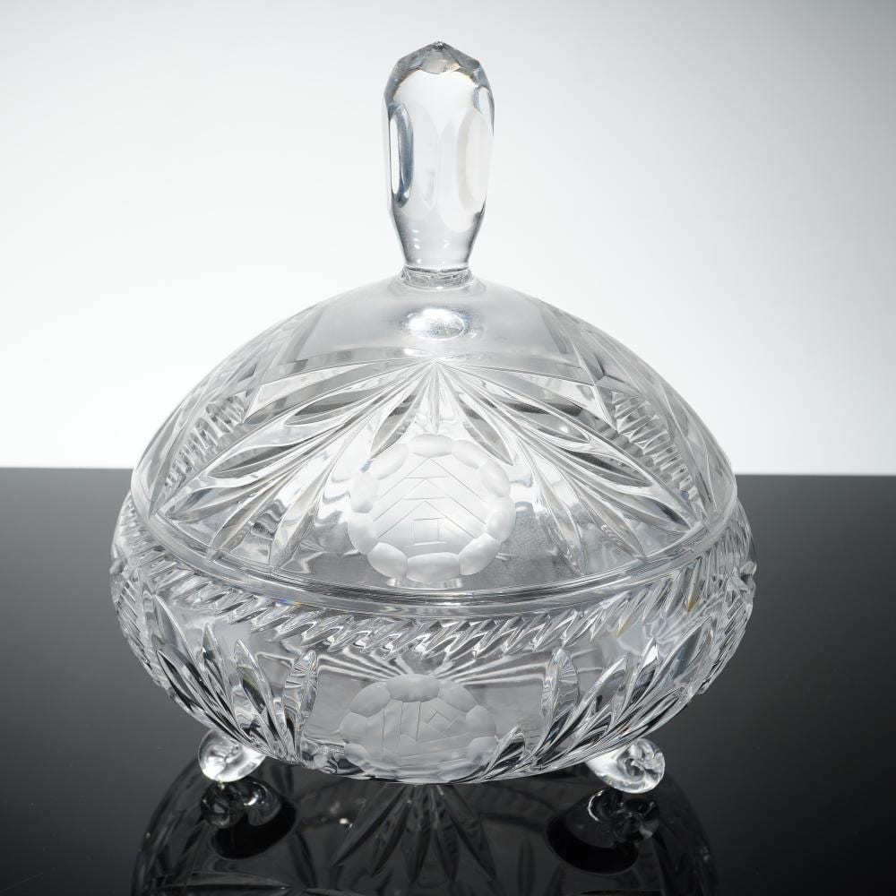 Clear glass footed candy dish with a faceted dome lid and decorative cut patterns, sitting on a dark reflective surface.