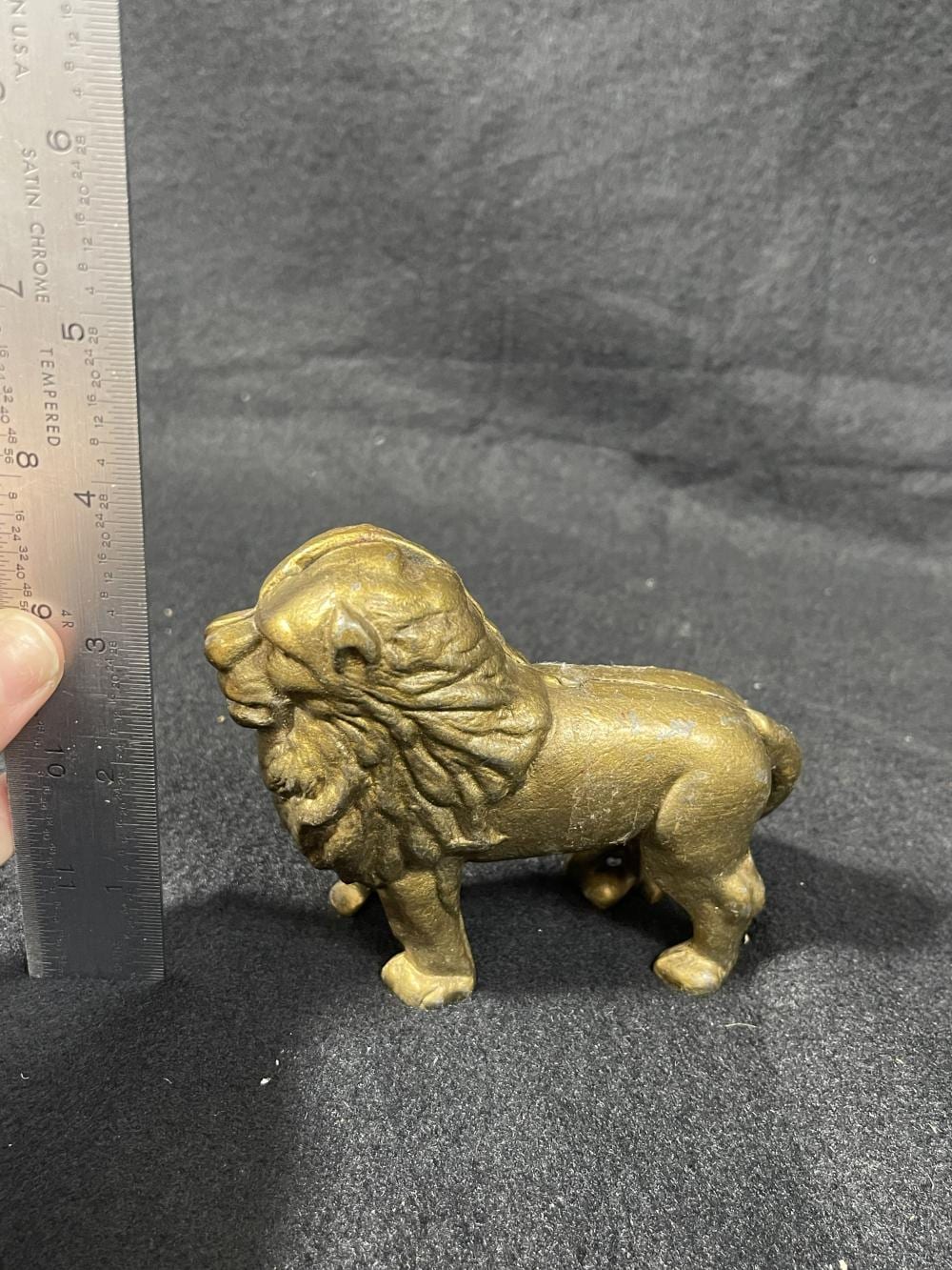 Small bronze-colored cast animal figurine shaped like a pig, shown side view on a gray surface with a ruler for scale.