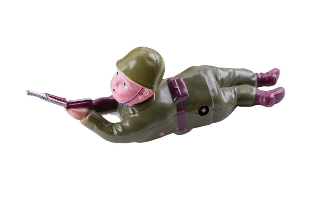 Mid-century toy soldier with a green helmet and uniform, pink face, holding a long silver rifle, lying in a posed position.
