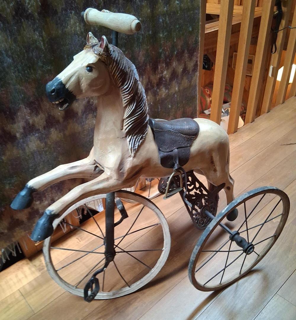 A carved wooden horse figure on a three-wheeled velocipede tricycle with a decorative saddle and wooden spoked wheels.