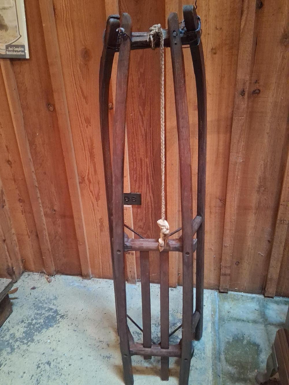 Antique-style wooden sled with curved runners and dark leather straps, standing indoors against a wooden wall.