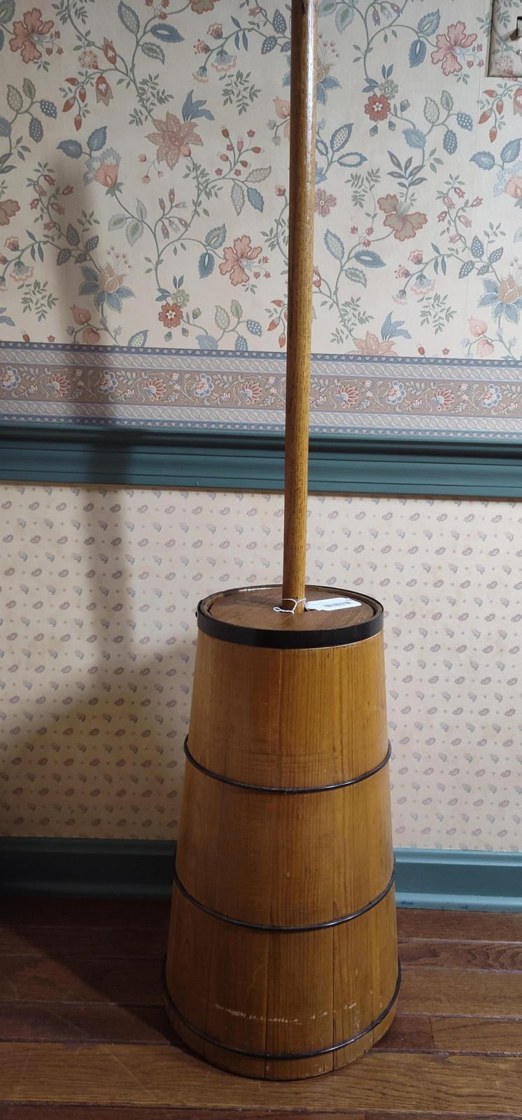 Antique-style wooden butter churn with a tall central dasher and a circular lid, standing on a floor in front of floral wallpaper.