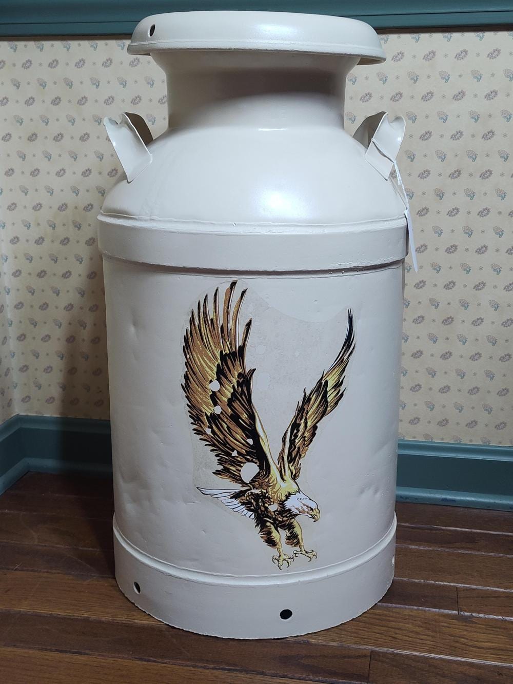 White metal milk can with a round body, handle on top, and a gold-and-black eagle graphic on the front.