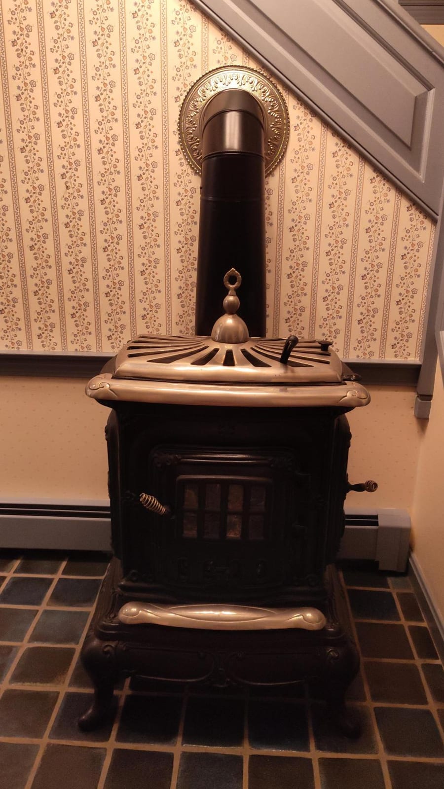 Antique-style black wood-burning parlor stove with brass top and ornamental front panel, standing in front of patterned wallpaper.