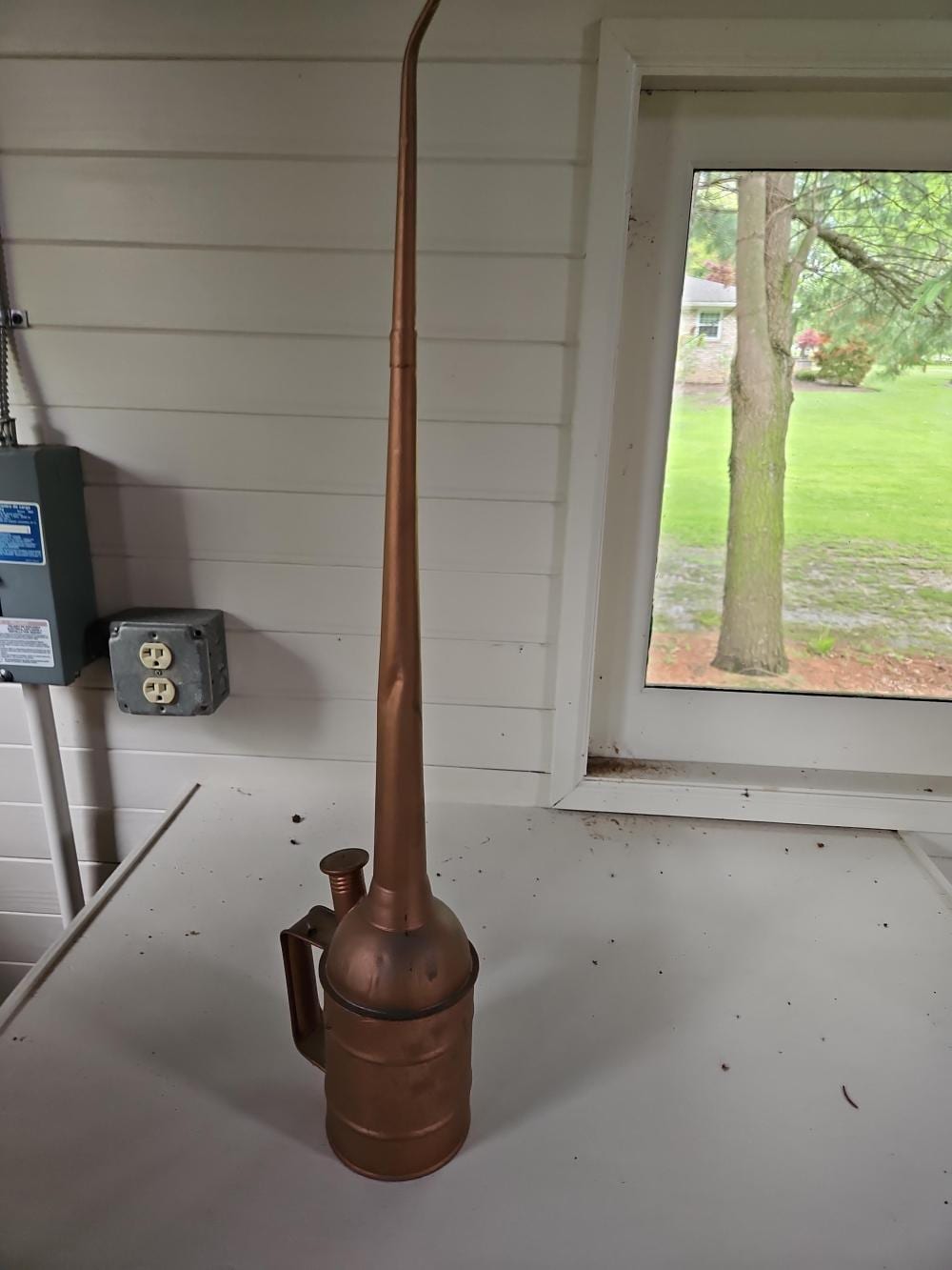 Copper-colored long-spout oil can standing indoors near a window.