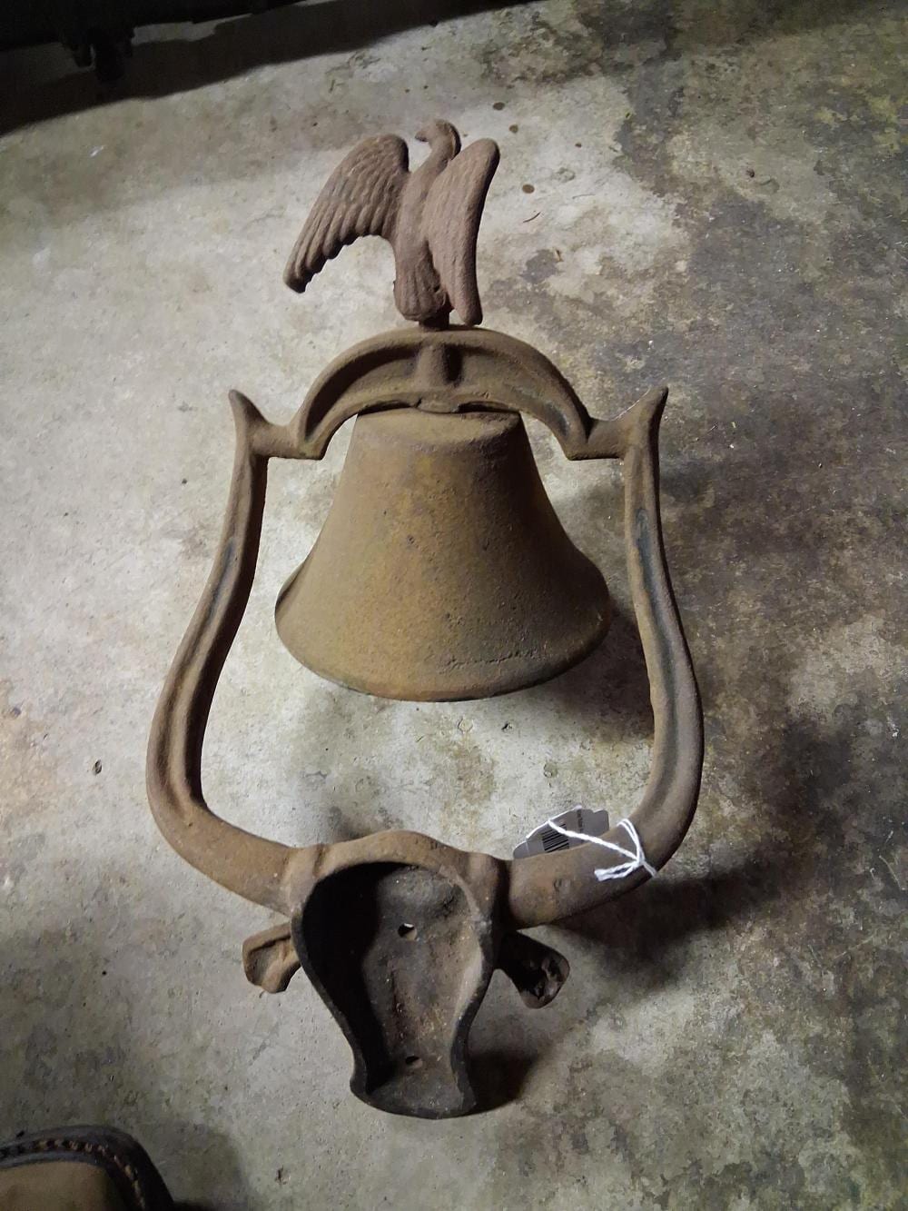 Cast iron dinner bell with a bell cup body and an eagle-like finial on top, shown against a gray floor.