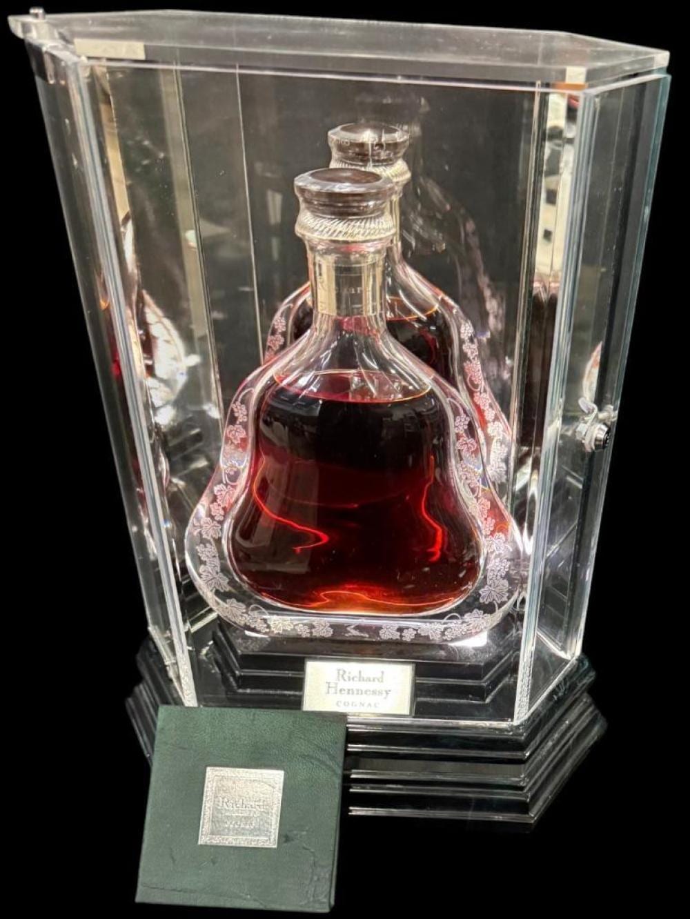 A dark glass cognac bottle with a decorative label inside a clear display case with a lit red interior glow; branded tags sit on the base.