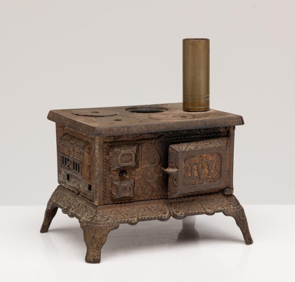 Cast iron miniature stove with ornate relief panels, four small legs, a round stovetop opening, and a cylindrical metal chimney pipe.