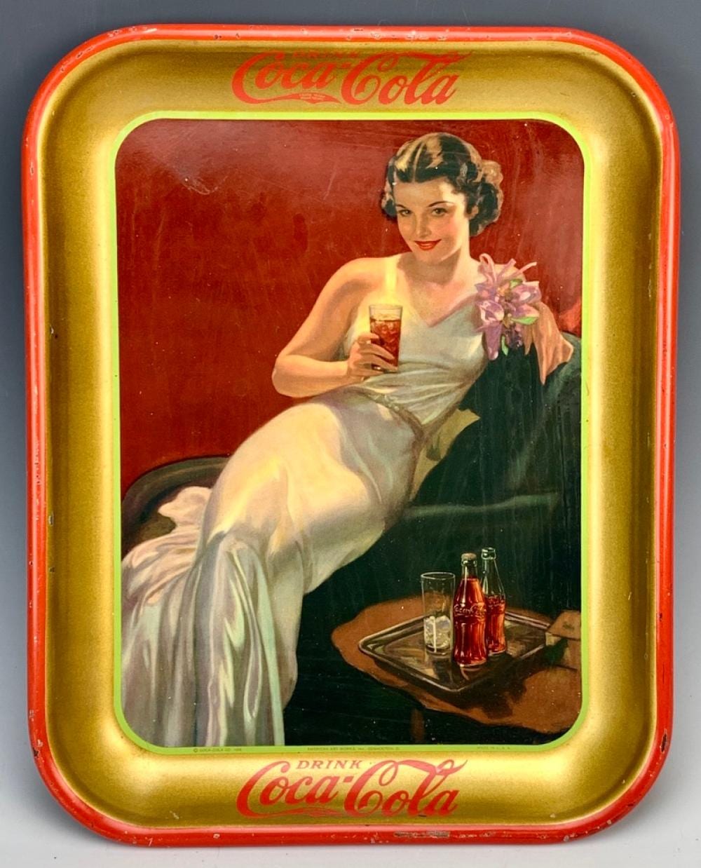 Vintage Coca-Cola metal tray with gold border and red sign text, featuring an illustration of a woman holding a glass bottle and flowers beside a drink table.