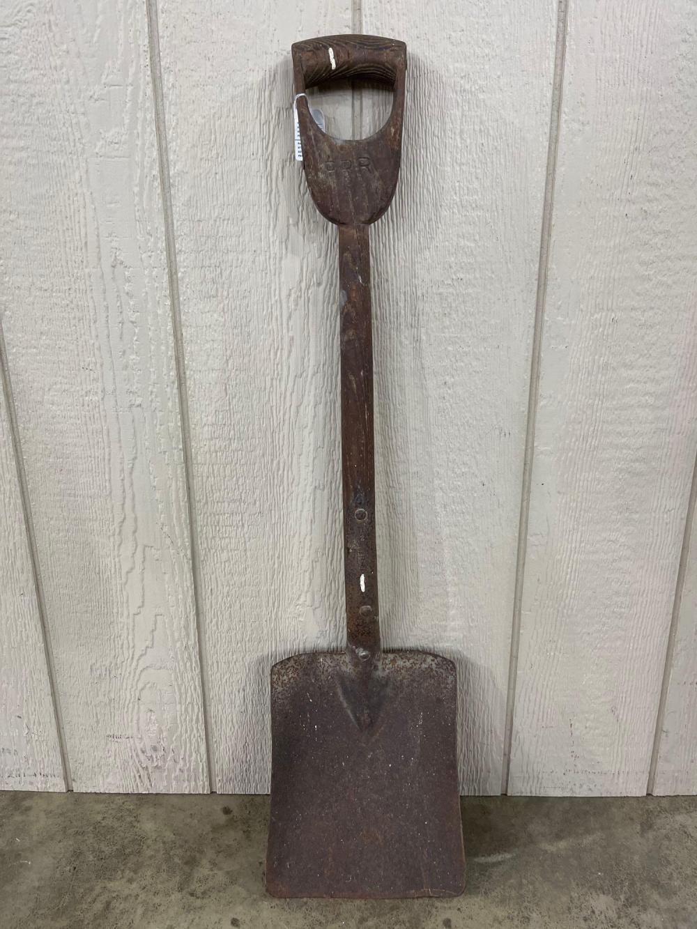 Brown metal coal shovel with D-shaped handle and a stamped marking near the top, standing upright against a light wall.