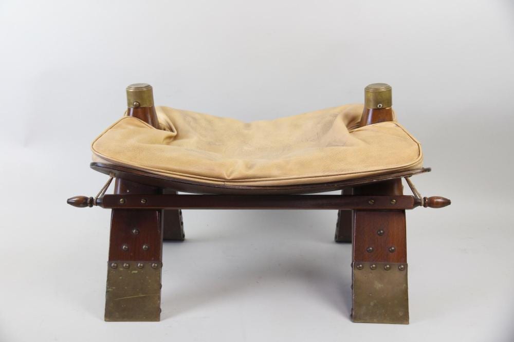 Brown wooden footstool with a curved frame and two short legs, topped by a light beige upholstered cushion and leather straps.