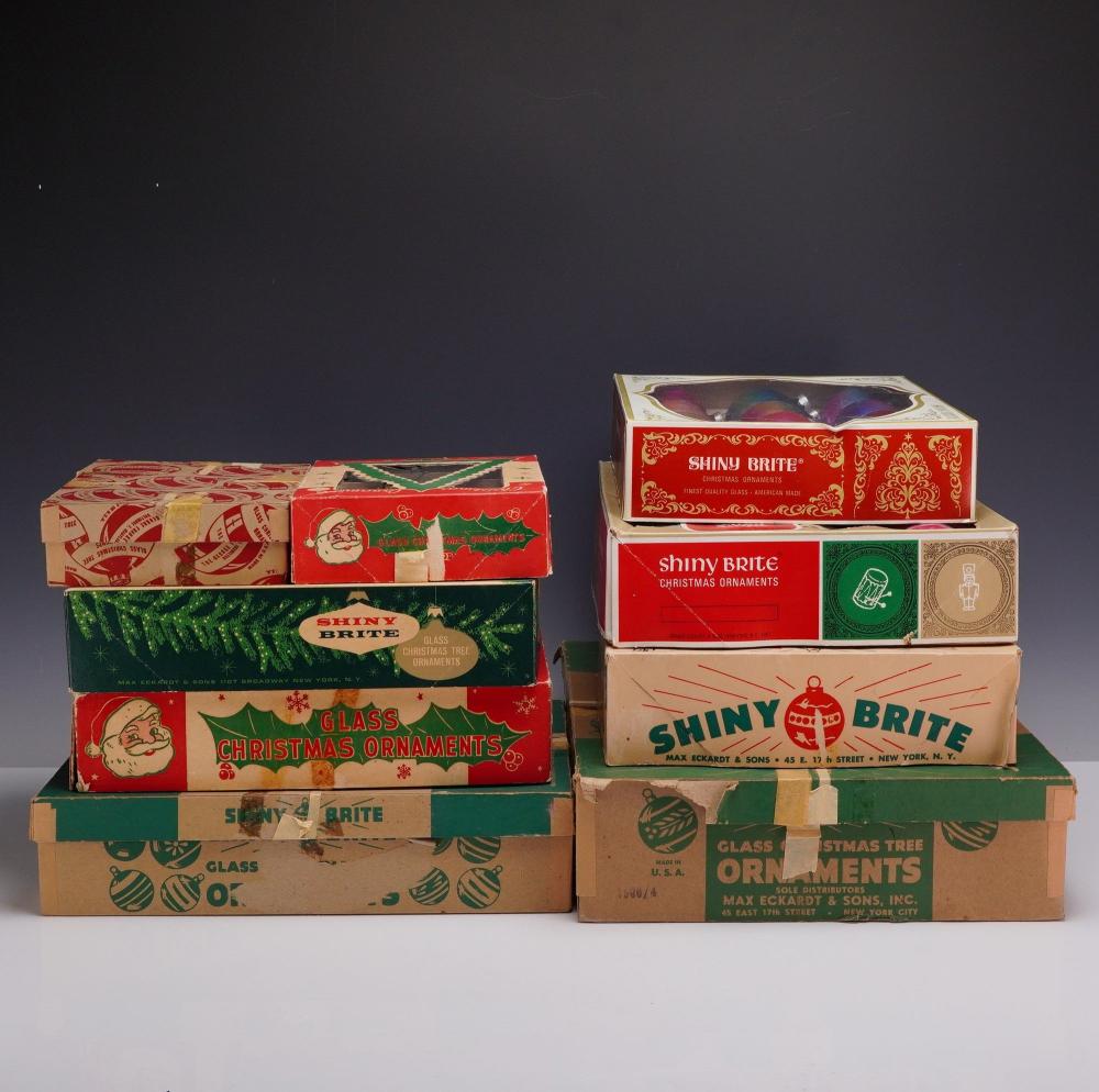 Stack of vintage Shiny Brite glass Christmas ornament boxes, red and green with Santa and ornament graphics, on a light background.