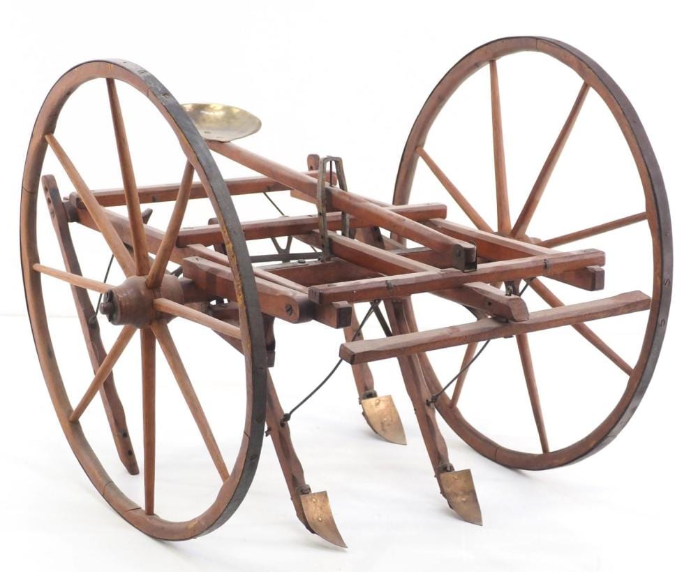 Wooden horse-drawn cultivator farm implement with two large spoked wheels, metal frame, and plow-like blades in front.