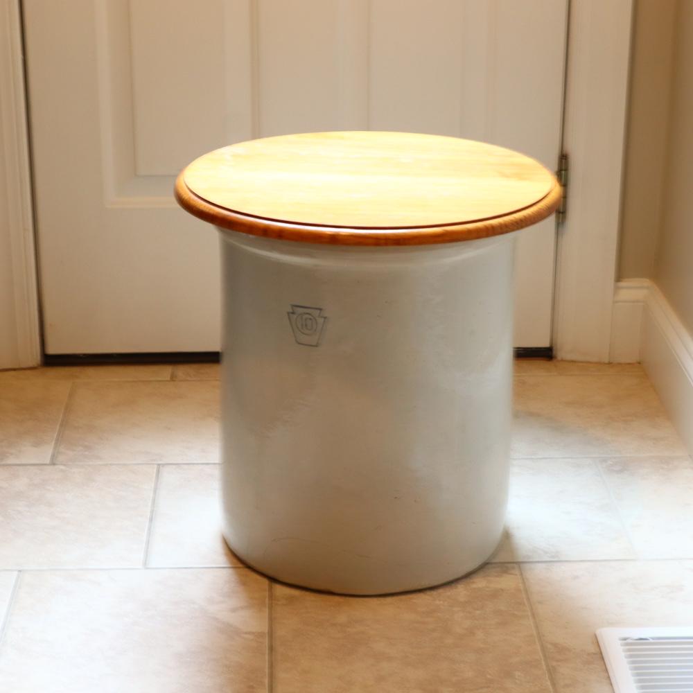 White stoneware crock with a round wooden lid and a small carved emblem on the front, photographed indoors on a tiled floor.