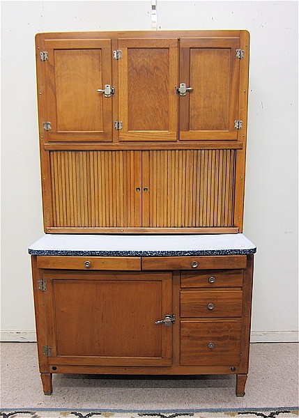 Wooden two-section Hoosier-style kitchen cabinet with doors, drawers, and hinged top compartments against a wall.