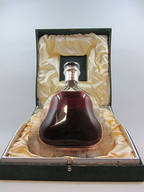 Glass cognac bottle with amber liquid and a metallic pour spout, set inside an open green presentation box lined with gold satin-like fabric.