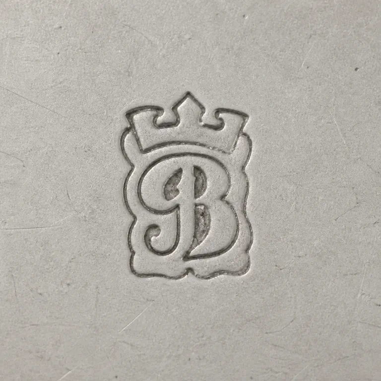 J B: Tiny ornate J-like/B-like monogram and its cartouche or crest silhouette; oxidized reconstruction.