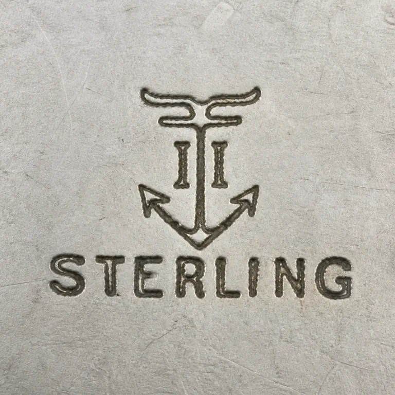 William Nost Co.: A compact anchor-like device with a flared top, two vertical strokes beside the shaft, and STERLING below; oxidized reconstruction.