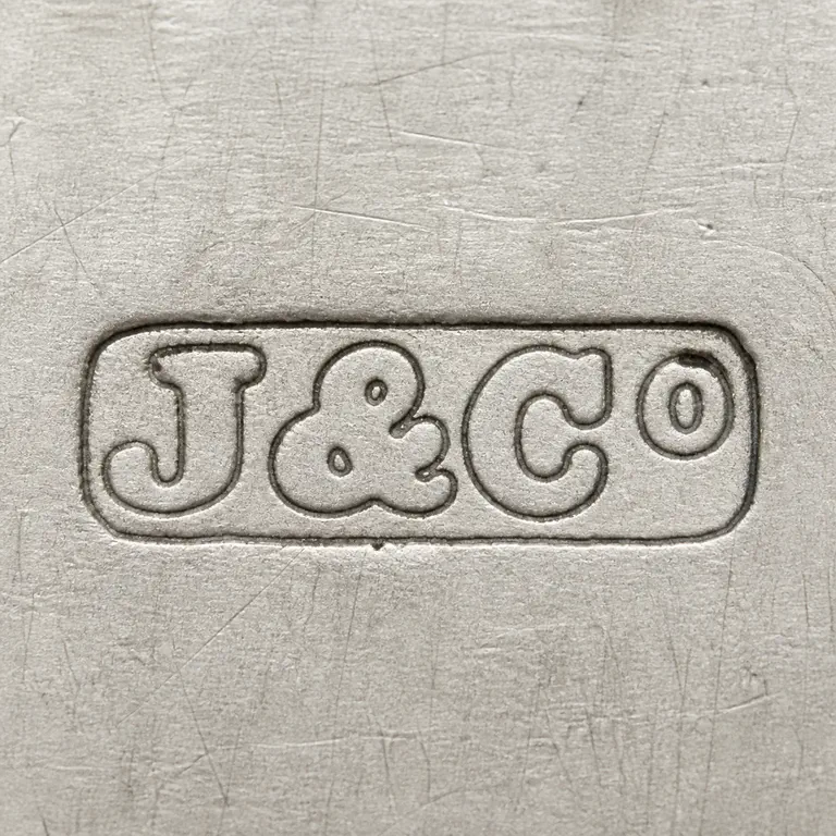Joseph Jennens & Co: J-like, ampersand-like, C-like, and small O-like terminal groups are retained left to right inside one worn rounded horizontal cartouche; oxidized reconstruction.