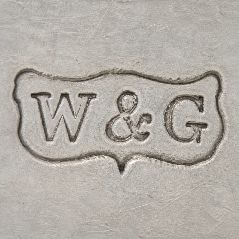 W & G: Long irregular plaque with W, ampersand-like central mark, and G-like form, rendered as unfilled contours; oxidized reconstruction.