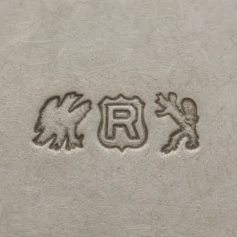 Reed & Barton: Three source-order groups: a worn left winged silhouette, uppercase R inside one scalloped shield, and a worn right upright silhouette; oxidized reconstruction.